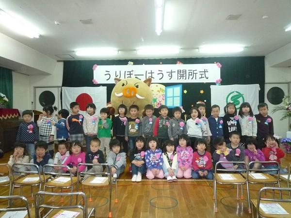 「うりぼーはうす 開所式」の横断幕の前で、園児達と「まるいの」が一緒に写っている集合写真